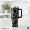 Heavy-duty black 'Stanley-style' insulated tumbler with a white heart print pattern, featuring a large ergonomic handle and clear straw, branded Beezmoon Code 127.