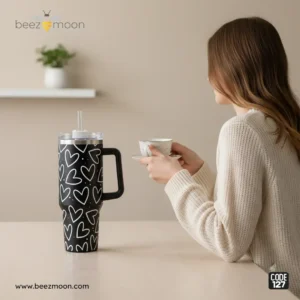 Heavy-duty black 'Stanley-style' insulated tumbler with a white heart print pattern, featuring a large ergonomic handle and clear straw, branded Beezmoon Code 127.